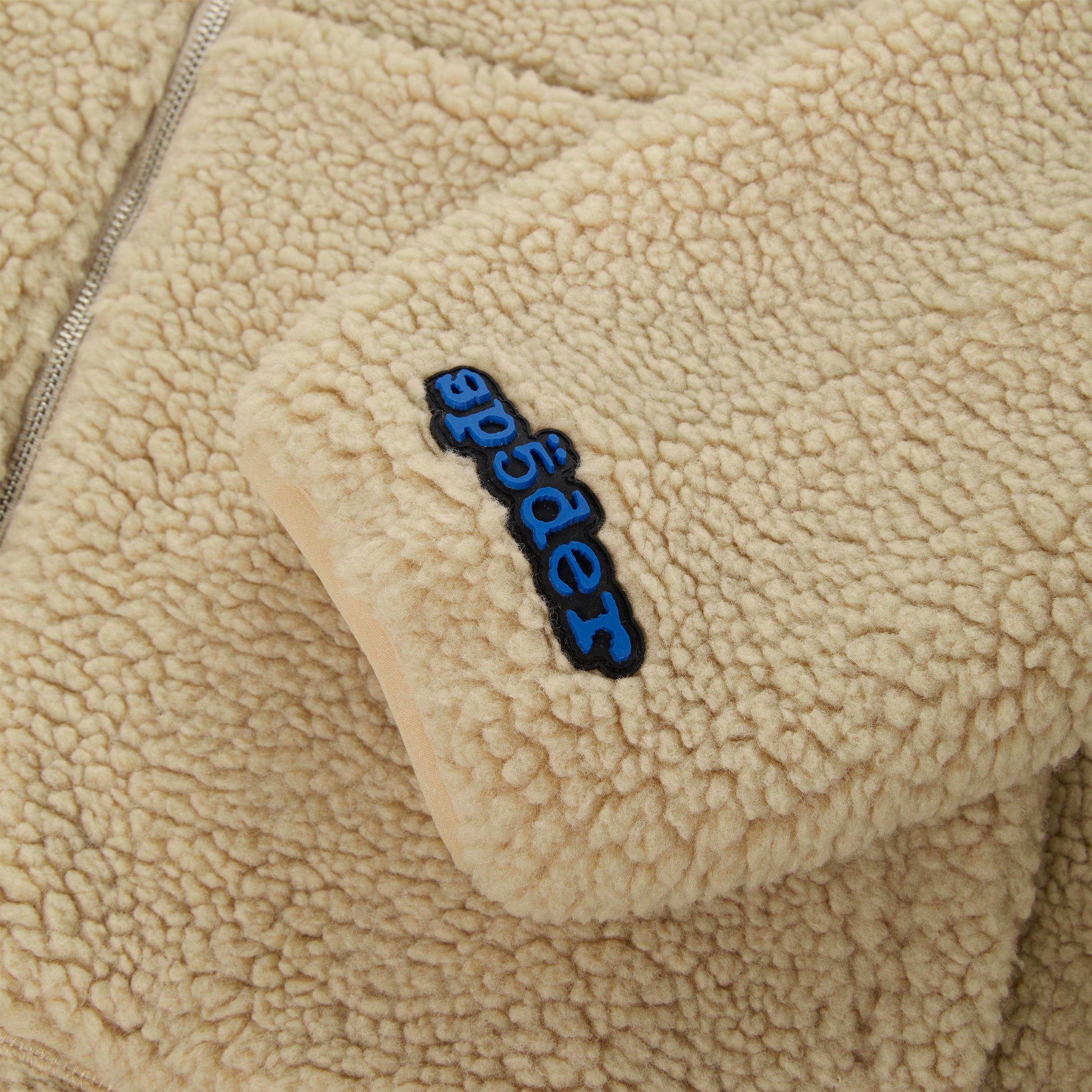 Detail view of rubberized logo patch on SP5DER Teddy Fleece Zip-Up in Brown